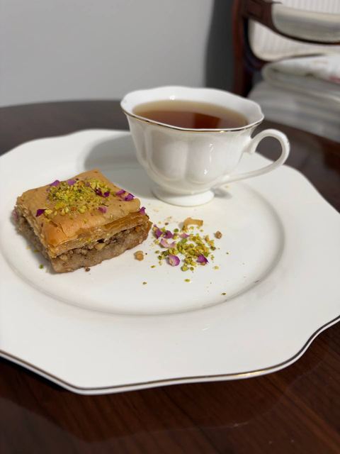 A square of pistachio baklava on a white scalloped plate beside a cup of tea