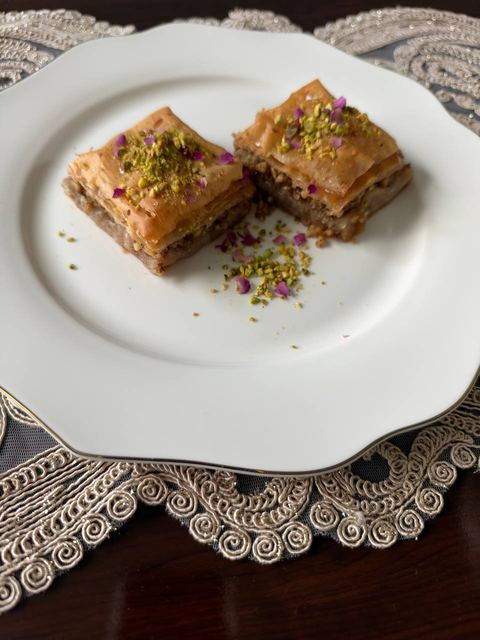Two pistachio baklava squares on a white plate with a lace placemat underneath